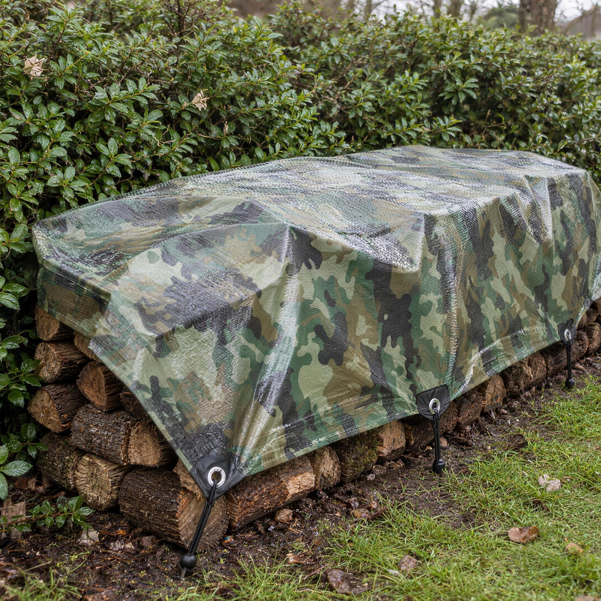 Camouflage tarpaulin covering a log pile against a garden hedge, tied with bungees