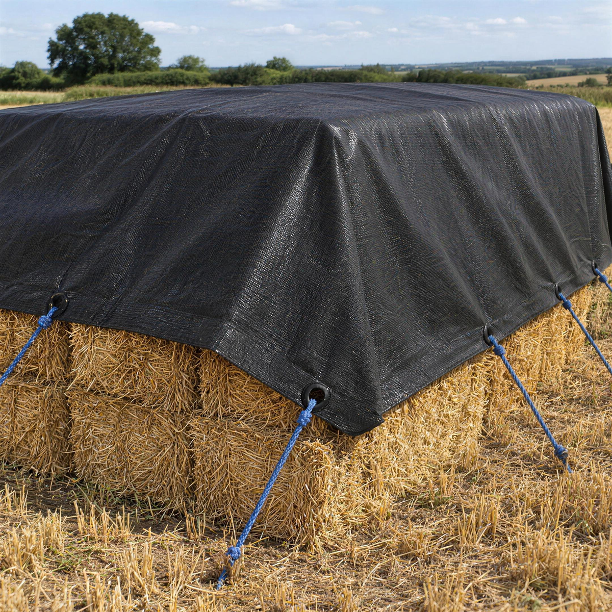 Black 200gsm tarpaulin covering a stack of straw bales in a stubble field, roped down through its plastic eyelets